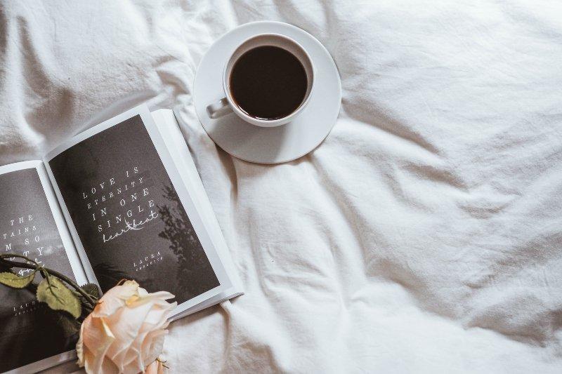 A photo of white bed sheets with a black cup of coffee, a book, and a rose